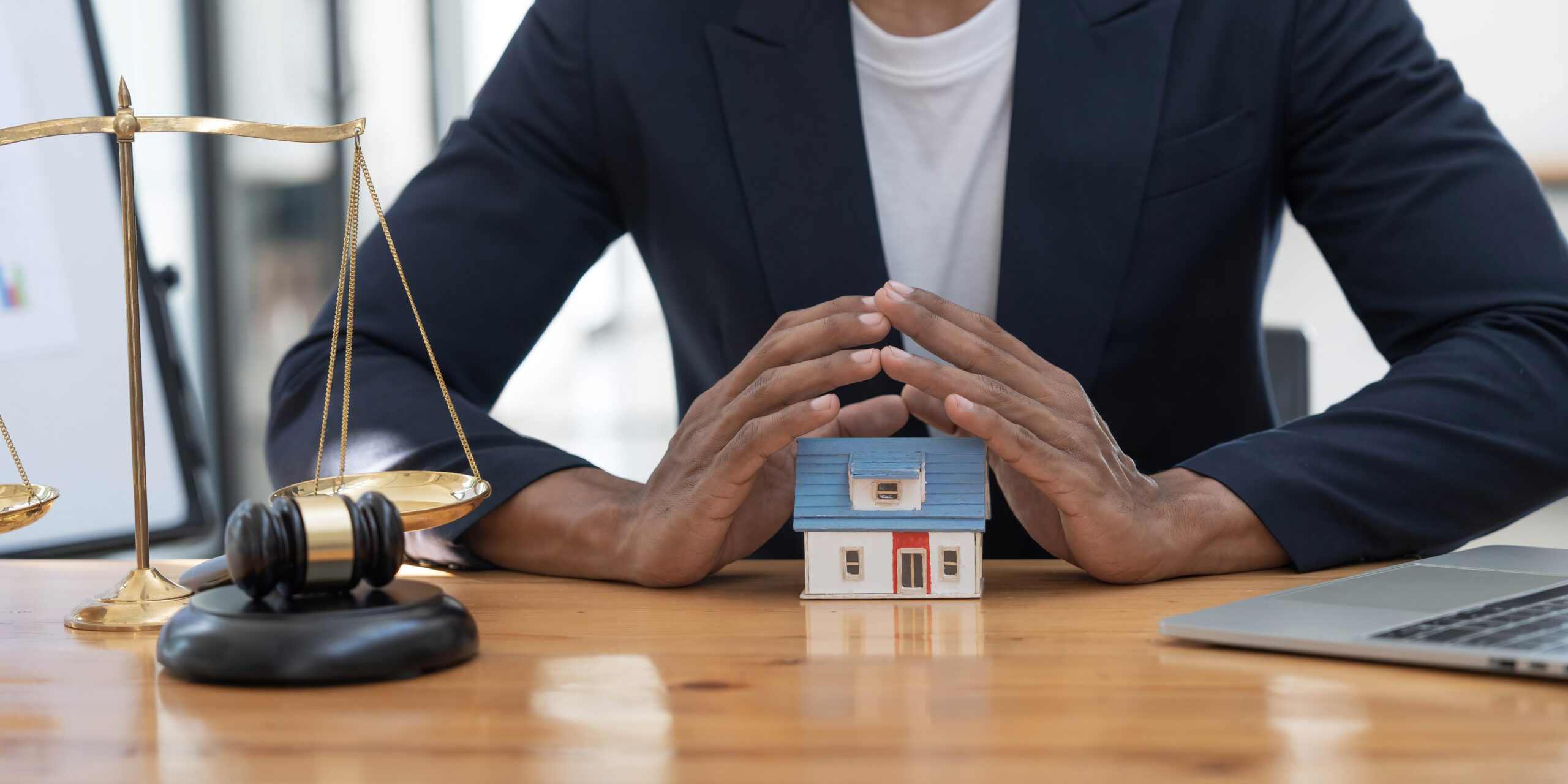 Close-up of a model house in front of a lawyer with a hammer and a laptop in an office.