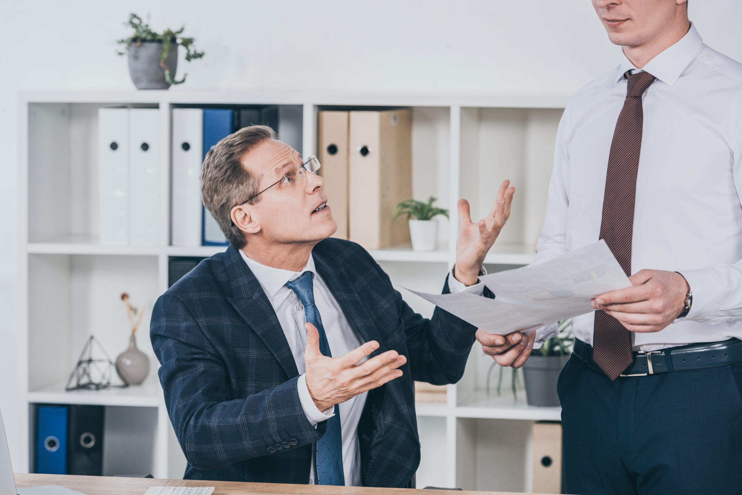 middle aged dissatisfied businessman sitting at table and talking with worker standing near with